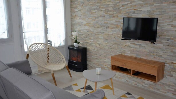 Una sala de estar con un sofá, una silla de mimbre, una mesita, un televisor montado en una pared de piedra y un mueble de madera.