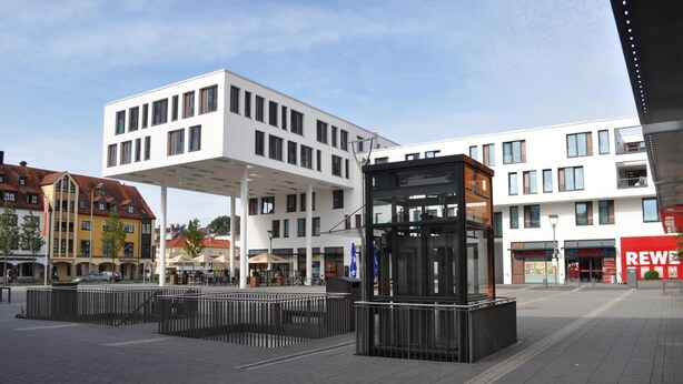 Una plaza urbana moderna con un área de asientos públicos y un quiosco con fachada de vidrio.