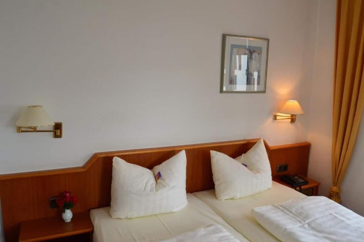 Habitación de hotel con dos camas, una mesita de noche con lámpara, un cuadro en la pared y un jarrón con flores.
