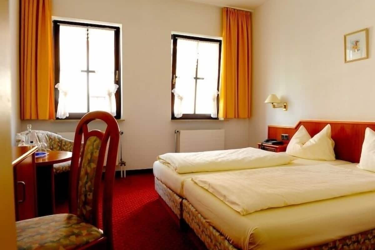Una habitación de hotel con una cama, un escritorio, una silla, dos ventanas con cortinas y una lámpara fijada a la pared.