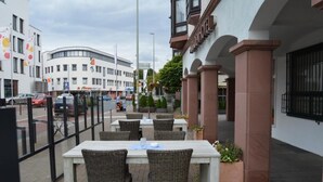 Un espacio al aire libre con mesas y sillas, una entrada con reja y un edificio de fachada blanca y pilares rojos.