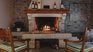A stone fireplace with a wooden mantel, two chairs with green cushions, and a potted plant. - Il Fontego (Borgo Valbelluna)