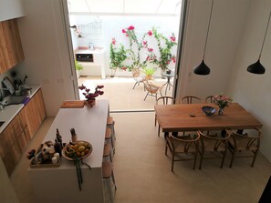 A modern kitchen with wooden cabinets, a dining area with a wooden table and chairs, and a view of a balcony with potted plants and flowers.