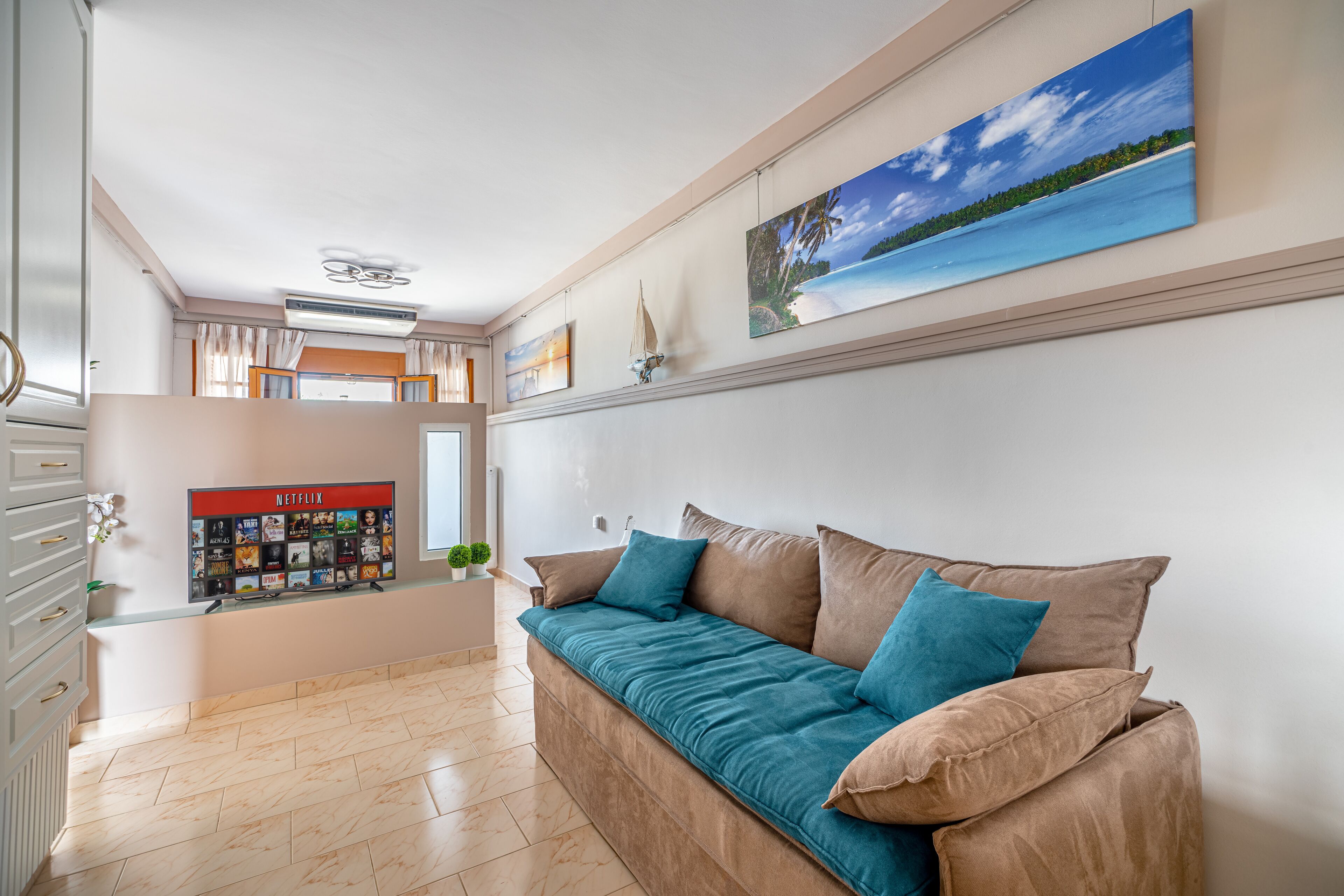 A modern living room with a beige sofa, teal cushions, a mounted artwork of a beach scene, and a wall-mounted Netflix display.