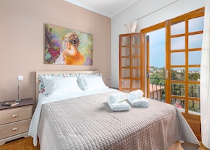 A bedroom with a large bed, a painting on the wall, and a view of the outside through an open window.