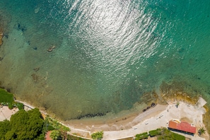 An aerial view of a coastal area with a beach, clear water, and a building with a red roof.