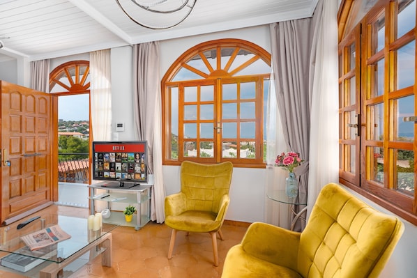 A room with a large window, a yellow armchair, a glass table with a newspaper, and a view of a town.
