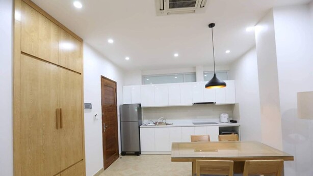 A compact kitchen with wooden cabinets, a stainless steel refrigerator, a dining table with chairs, and recessed ceiling lighting.