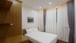 A bedroom with a bed, a wooden nightstand, a wall-mounted shelf, and a window with sheer curtains.