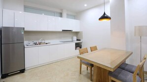 A modern kitchen with white cabinets, a stainless steel refrigerator, a wooden dining table with chairs, and a pendant light.