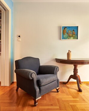 A room with a dark grey armchair, a wooden table with a vase, and a painting on the wall.