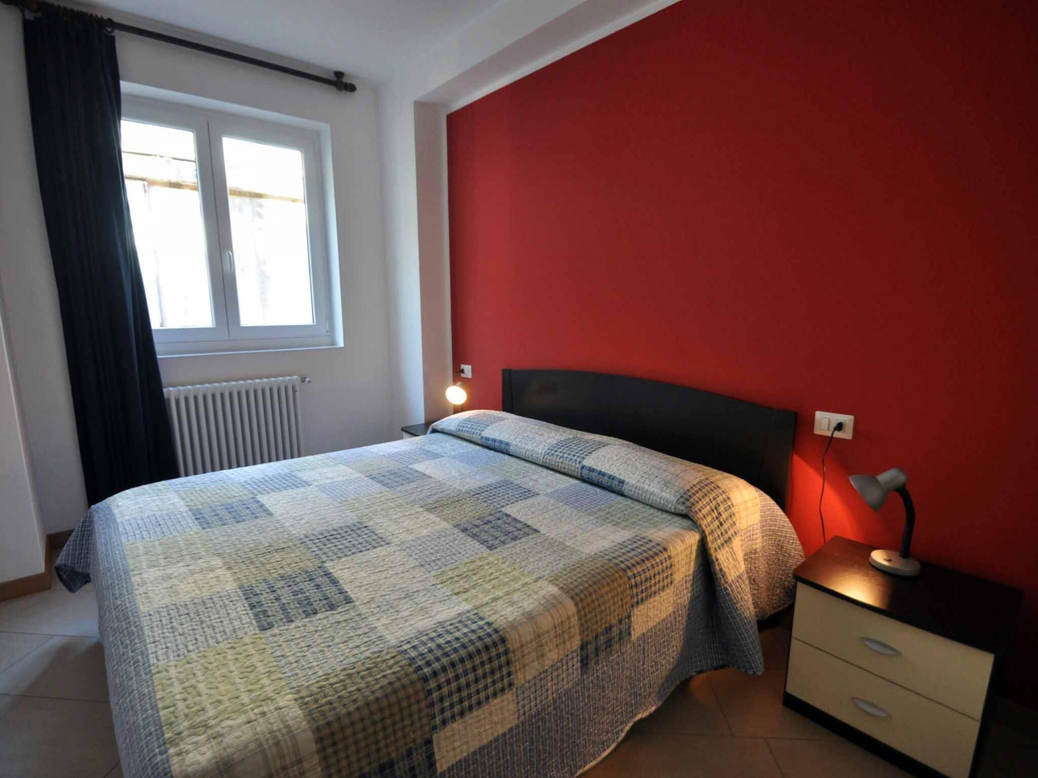 A bedroom with a bed, a nightstand, a lamp, a window with curtains, and a radiator.