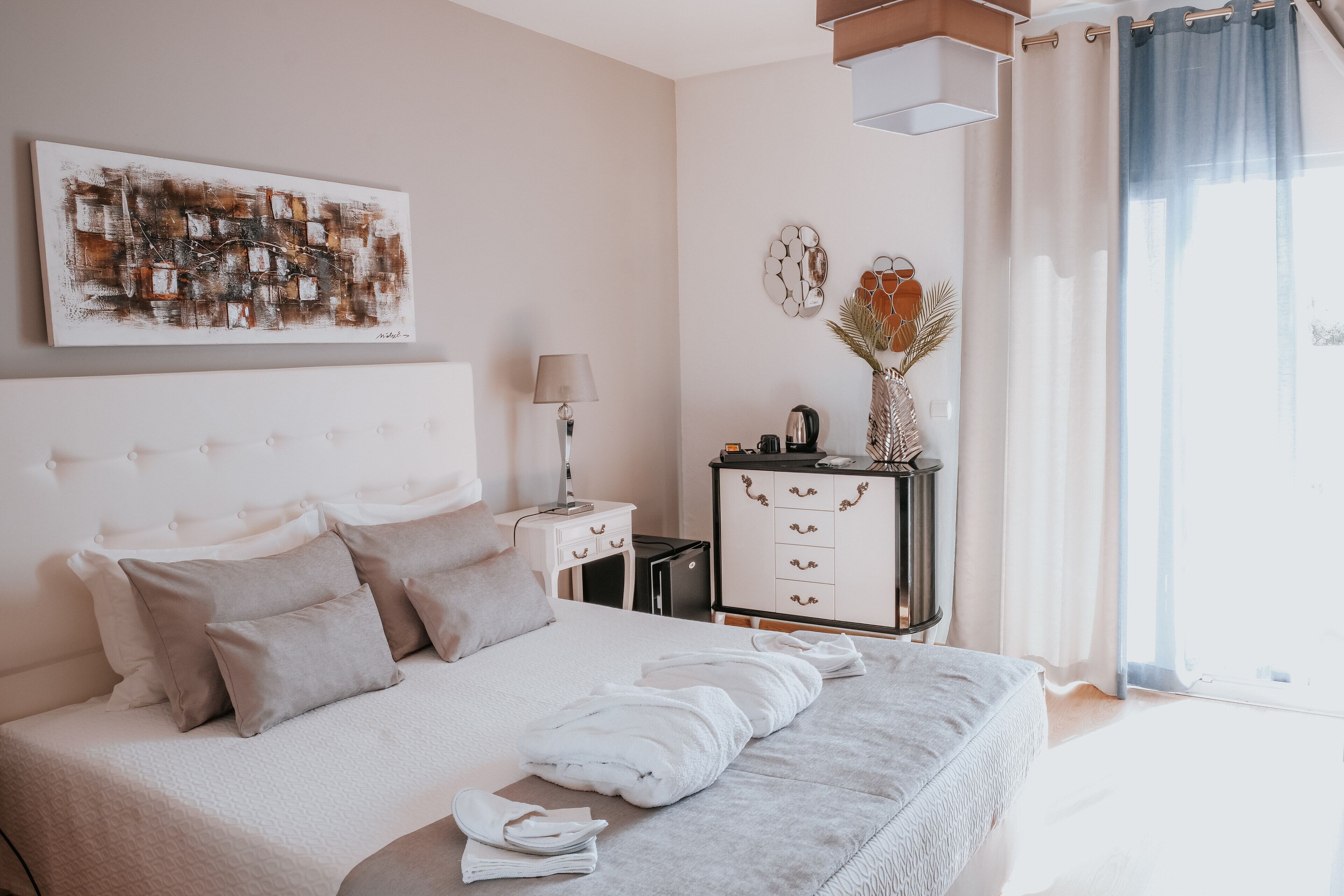 A modern bedroom with a large bed, a bedside table, a lamp, a dresser, and a painting on the wall.