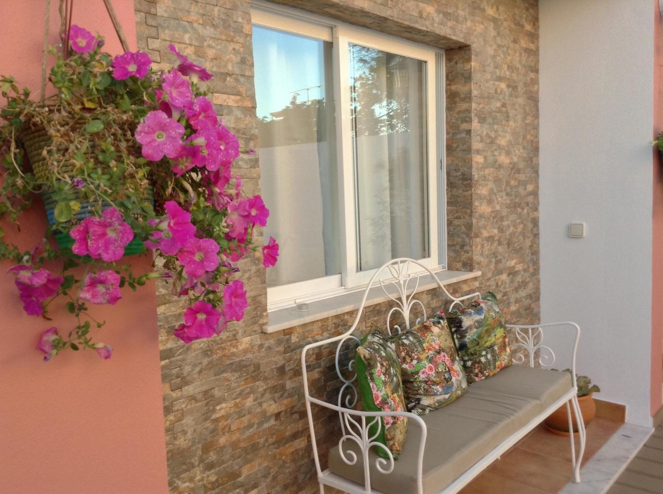 A stone wall with a window, a white metal bench with cushions, and hanging pink flowers.