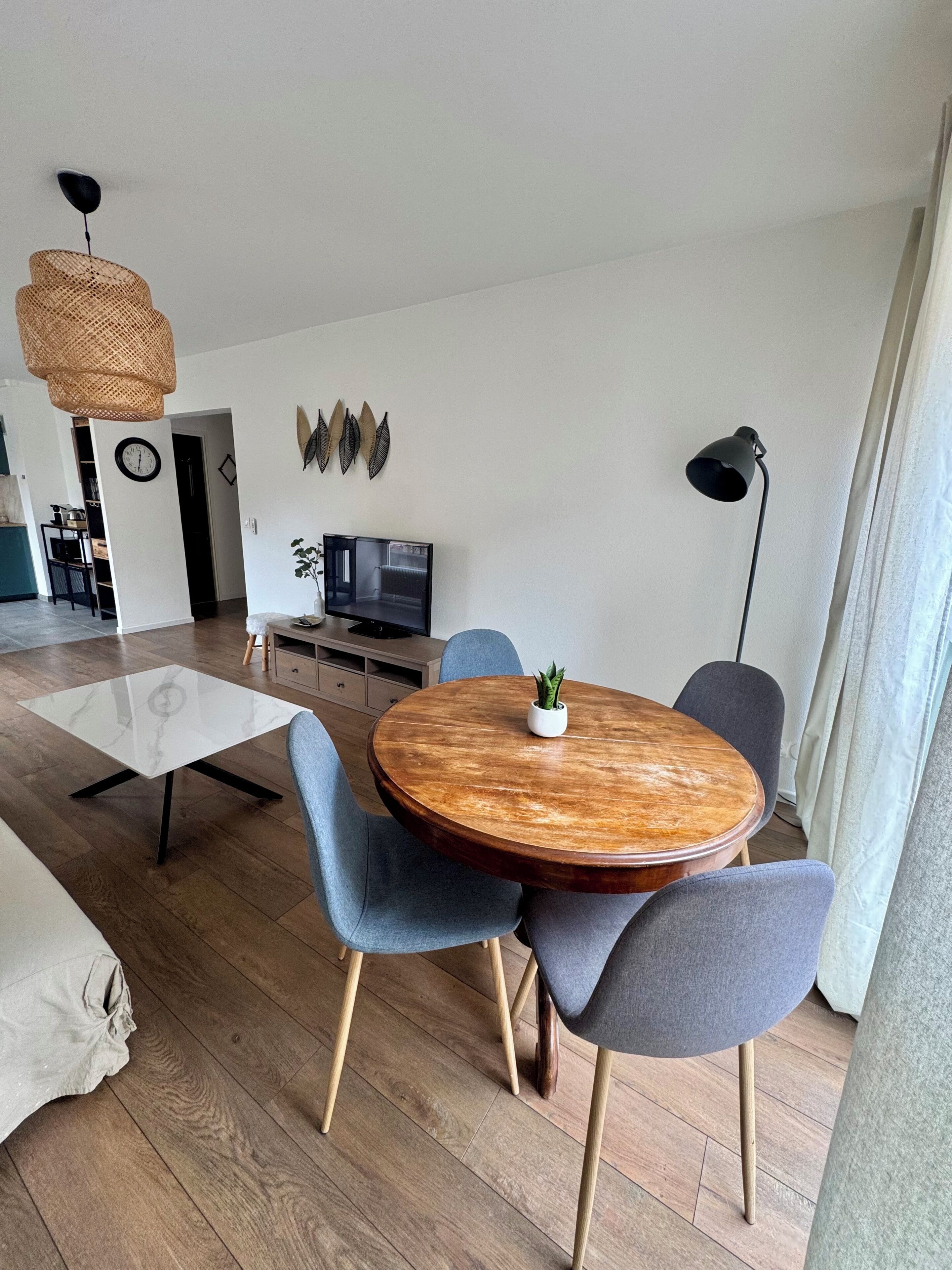 A modern living room with a round wooden dining table, two blue chairs, a white coffee table, a TV, a plant, and a hanging light fixture.