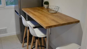Uma cozinha compacta com uma mesa de madeira, duas cadeiras brancas, uma geladeira de aço inoxidável e um pequeno vaso com planta.