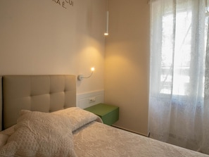 Un dormitorio con cabecera capitonada, una mesita de noche, una mesita auxiliar verde y una ventana con cortinas de encaje.