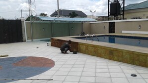 Una piscina con un borde empedrado, una persona agachada cerca y un edificio al fondo.