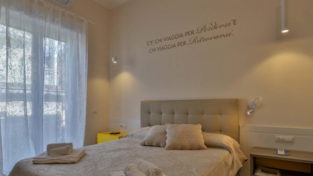 Una cama bien hecha con un cabecero color beige, una mesita de noche con una lámpara y una pared con una frase decorativa.