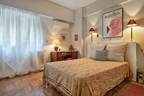 Un dormitorio con cama, lámparas de noche, aire acondicionado instalado en la pared, un cuadro enmarcado y una ventana con cortinas translúcidas.