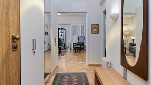 A hallway with a wooden door, a bench, a patterned rug, a mirror, and a view into a living area with a chair and a lamp.