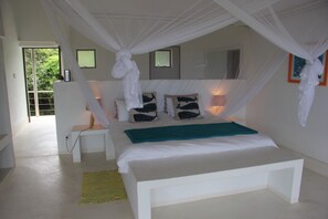 A bedroom with a canopy bed, a bedside table, a chair, a bedside lamp, and a view of the outdoors.