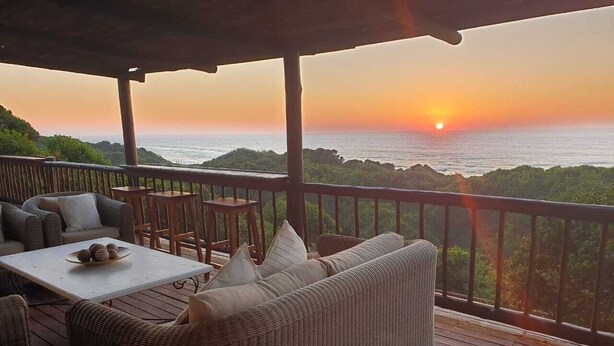 A wooden deck with wicker furniture and a table, overlooking a sunset over the ocean. - Kangela 11 (Ponta d'Ouro)