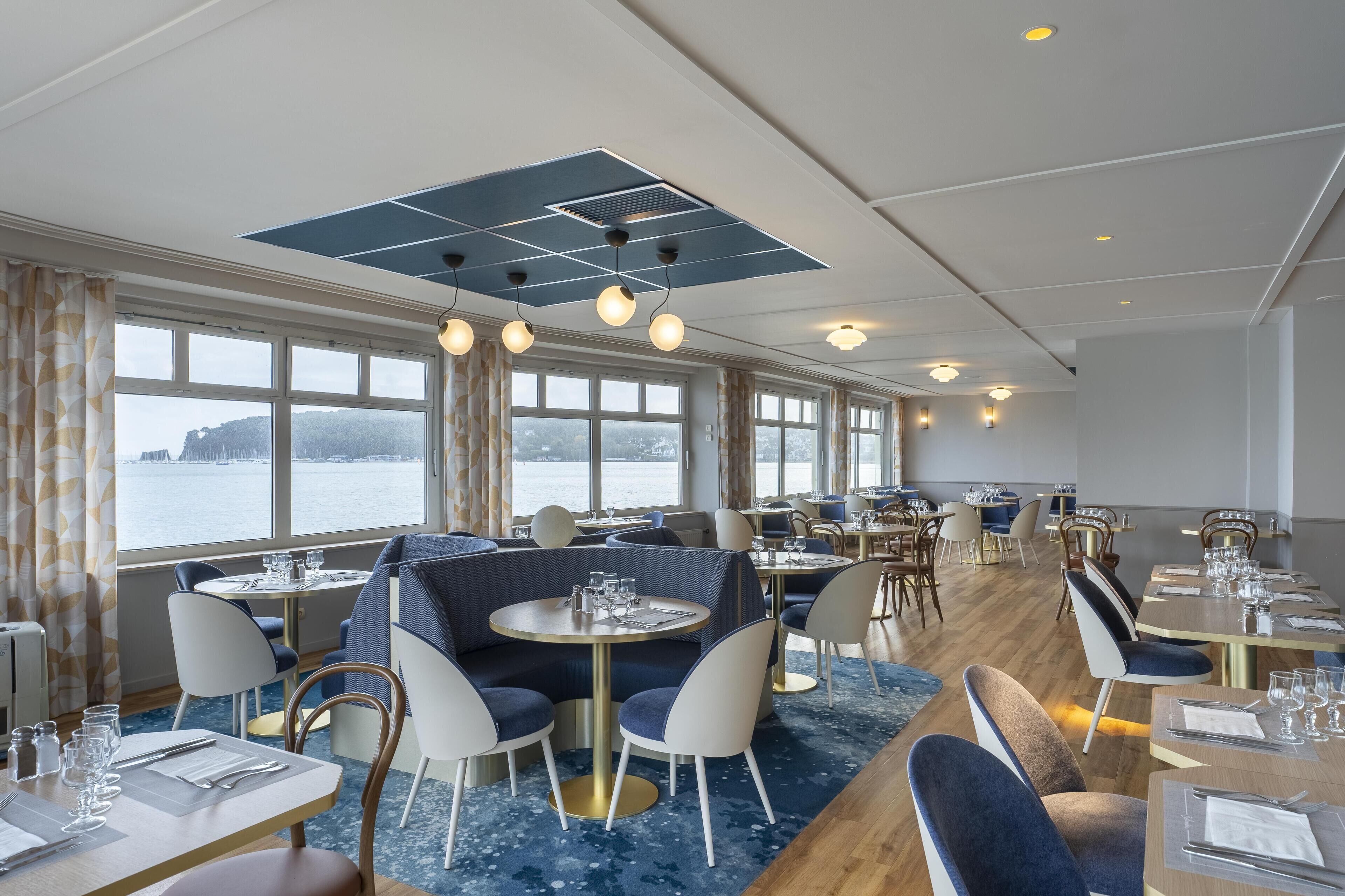 A restaurant with round tables, blue upholstered seating, and large windows overlooking a body of water.