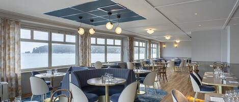En restaurang med runda bord, blå klädda sittplatser och stora fönster med utsikt över en sjö eller ett annat vattendrag.