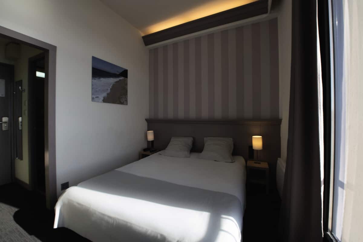 A hotel room with a large bed, two bedside tables with lamps, a headboard with vertical stripes, a framed picture on the wall, and a door to the left.