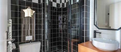 A modern bathroom with a black and white checkered tile wall, a glass-enclosed shower, a white toilet, and a large oval sink with a mirror above it.