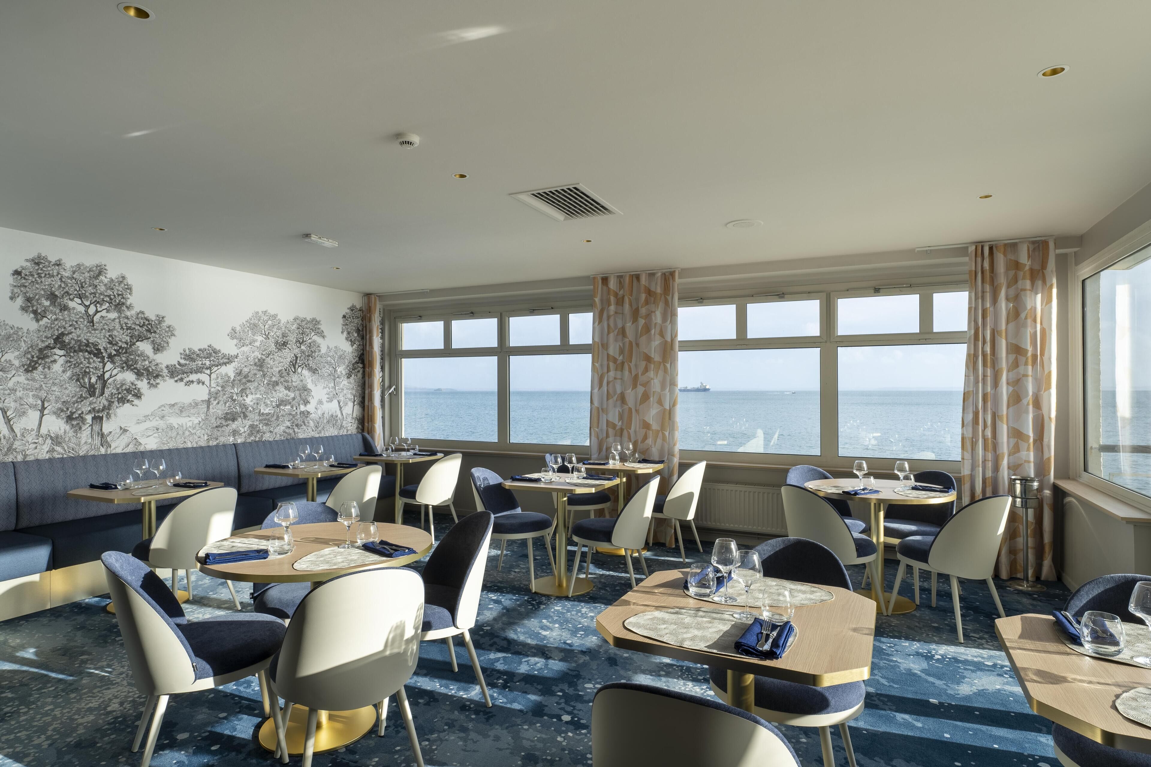 A dining area with tables and chairs, large windows with curtains, and a mural of trees on the wall.