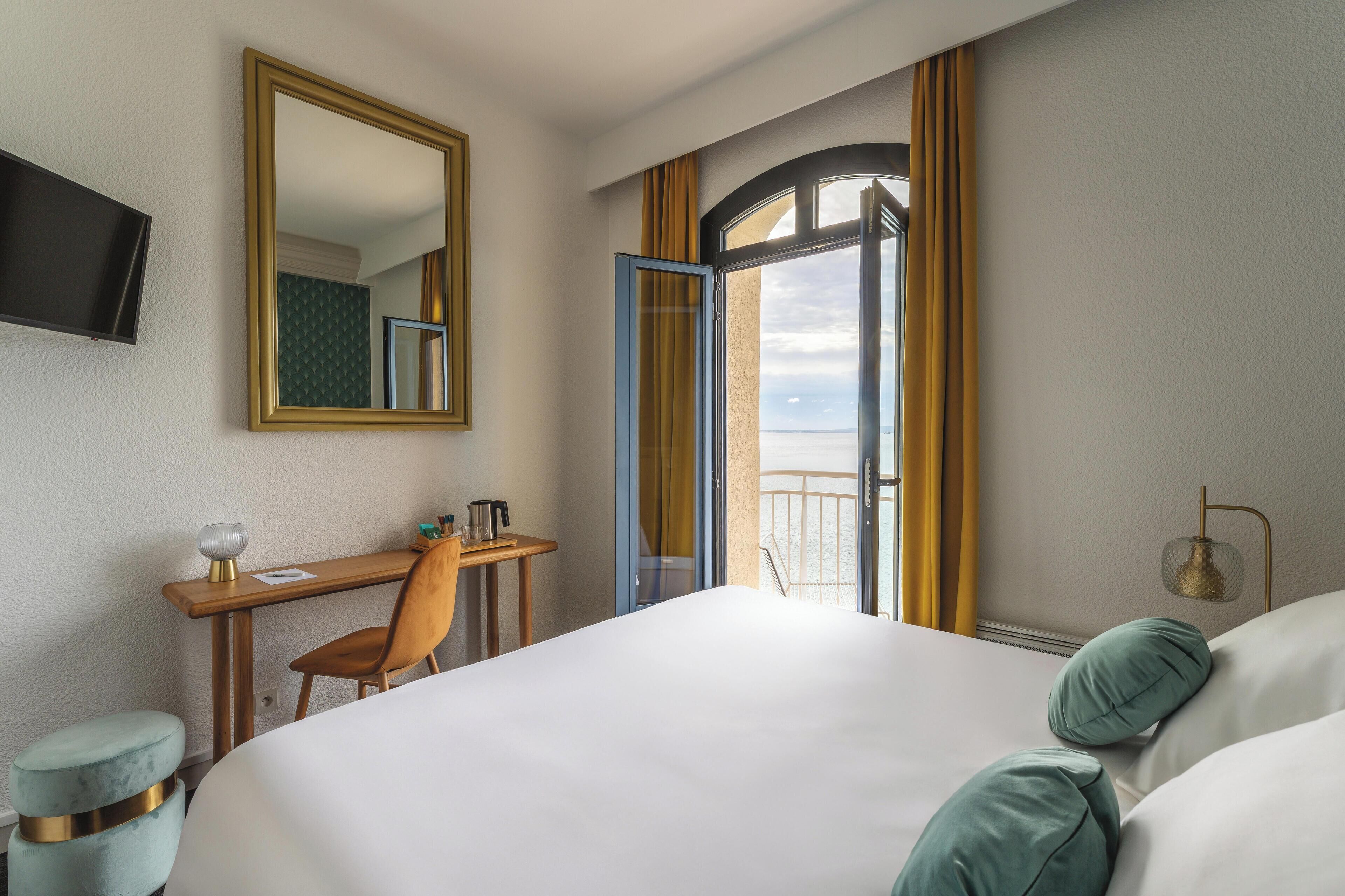A hotel room with a bed, a desk with a chair, a television, and a view of the sea through an open door.