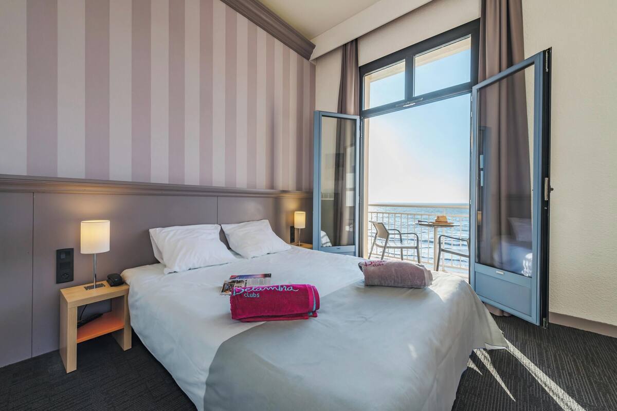 A hotel room with a bed, bedside table, lamp, and a view of the sea through an open door.