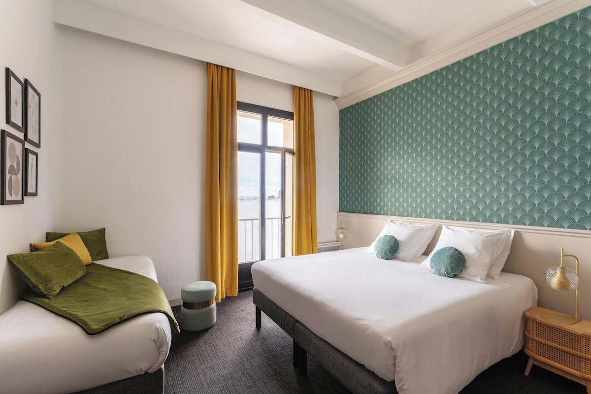 A hotel room with a bed, a sofa, a window with yellow curtains, a wall with a textured pattern, and a small side table with a lamp.