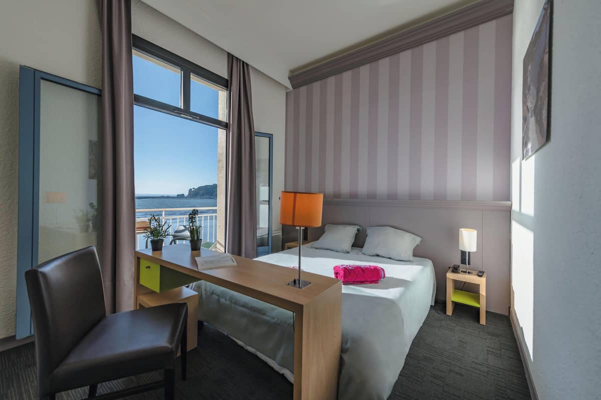 A hotel room with a bed, desk, chair, and a view of the sea.