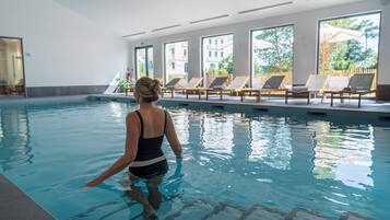 Une piscine intérieure à l’eau claire, baignée de lumière grâce à de grandes fenêtres, et bordée de chaises longues.