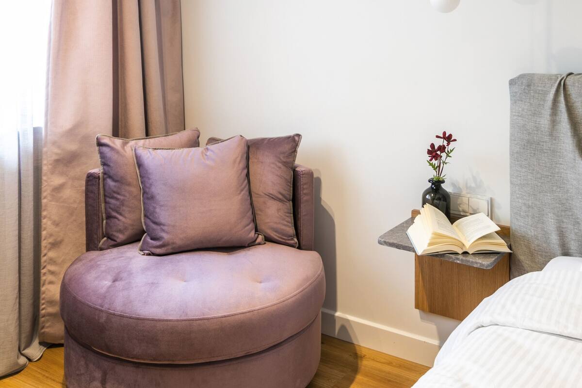 A modern bedroom with a round, purple upholstered chair, a bedside table with an open book, and a hanging light fixture.