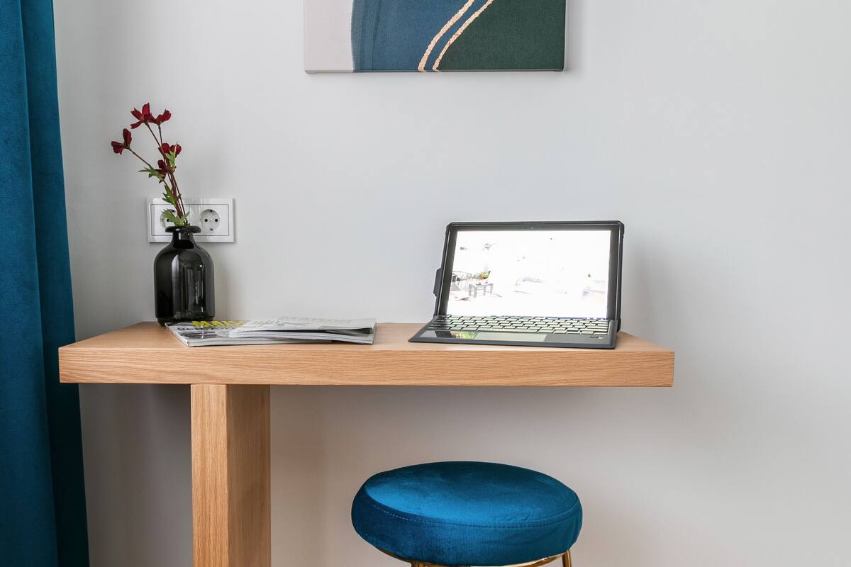 A wooden desk with a laptop, a vase with flowers, and a blue stool.