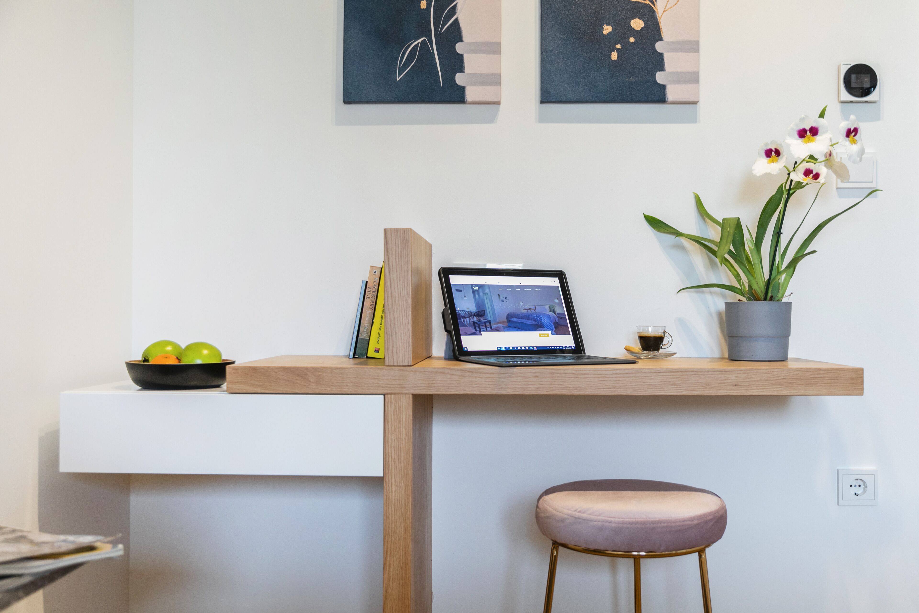 Um espaço de trabalho moderno com um laptop, livros, uma tigela de frutas, uma orquídea em vaso e uma cadeira minimalista.
