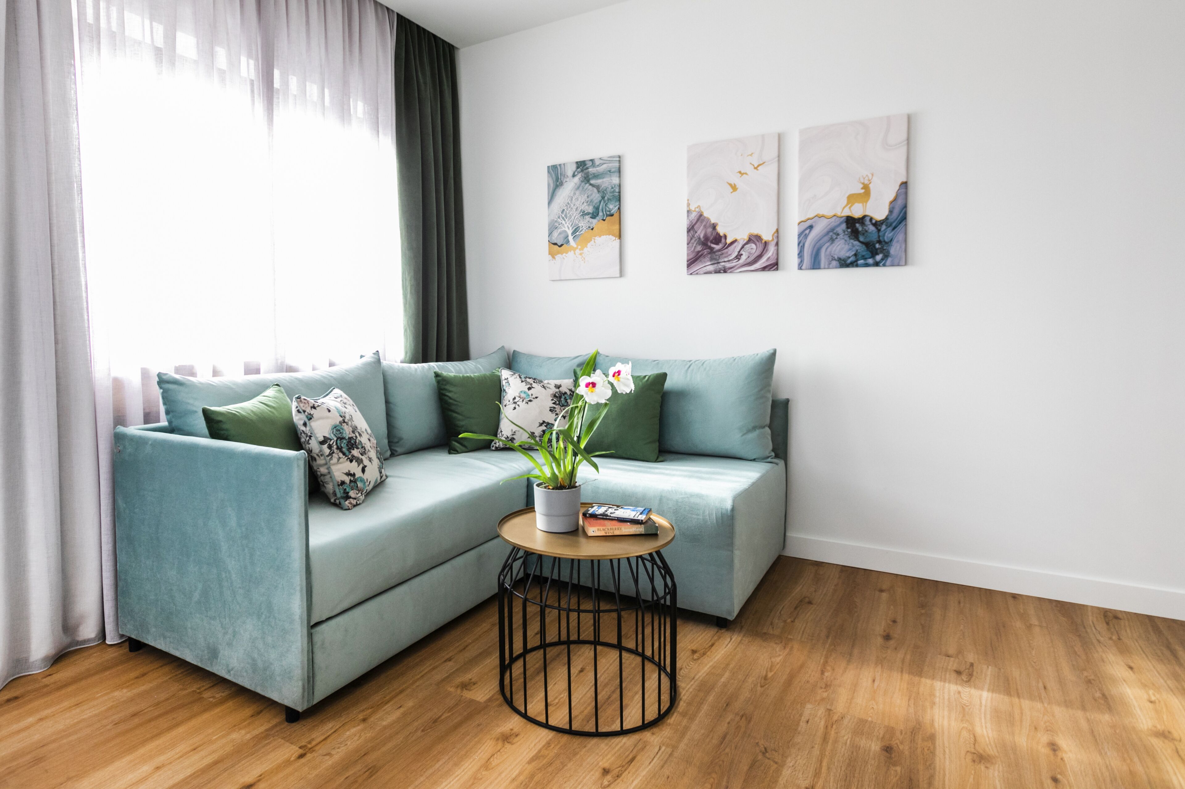 A modern living room with a teal sofa, green cushions, a round coffee table with a plant, and artwork on the wall.