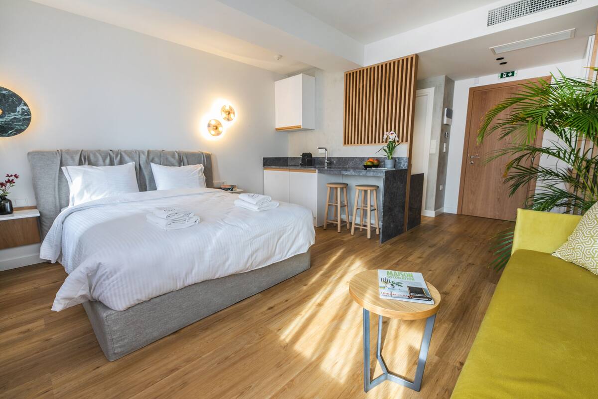 A modern hotel room with a large bed, a small kitchen area, a sitting area with a yellow sofa, and a wooden floor.