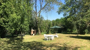 A grassy outdoor area with a wooden playhouse, a table with chairs, and a green umbrella. - GITE AUX FLEURS DE CERISES - Le Plessis Luzarches (95) _ 4 people (Le Plessis-Luzarches)