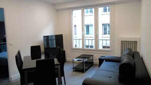 Un salon moderne avec un canapé noir, une table à manger avec des chaises, un téléviseur à écran plat et de grandes fenêtres donnant sur des immeubles.