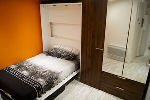 A single bed with a cityscape bedspread, a wooden wardrobe, and a mirror cabinet in a room with an orange accent wall.