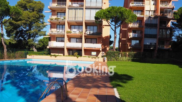 A swimming pool with a tiled deck, surrounded by a well-maintained lawn and a multi-story building with balconies. - COZY APARTMENT WITH AIR CONDITIONING & COMMUNAL SWIMMING POOL IN LOS PINOS (Platja d'Aro)