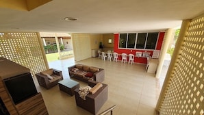 Ein geräumiger Wohnbereich mit einer Sofagarnitur, einem Couchtisch und einem Fernseher. Es gibt einen Balkon mit Stühlen und einem Tisch sowie eine Küchenzeile mit roter Wand und großen Fenstern.