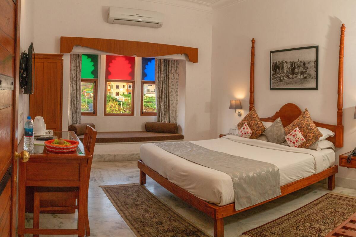 A bedroom with a four-poster bed, a wooden desk, a chair, a bedside table, a lamp, and a window with colorful shutters.