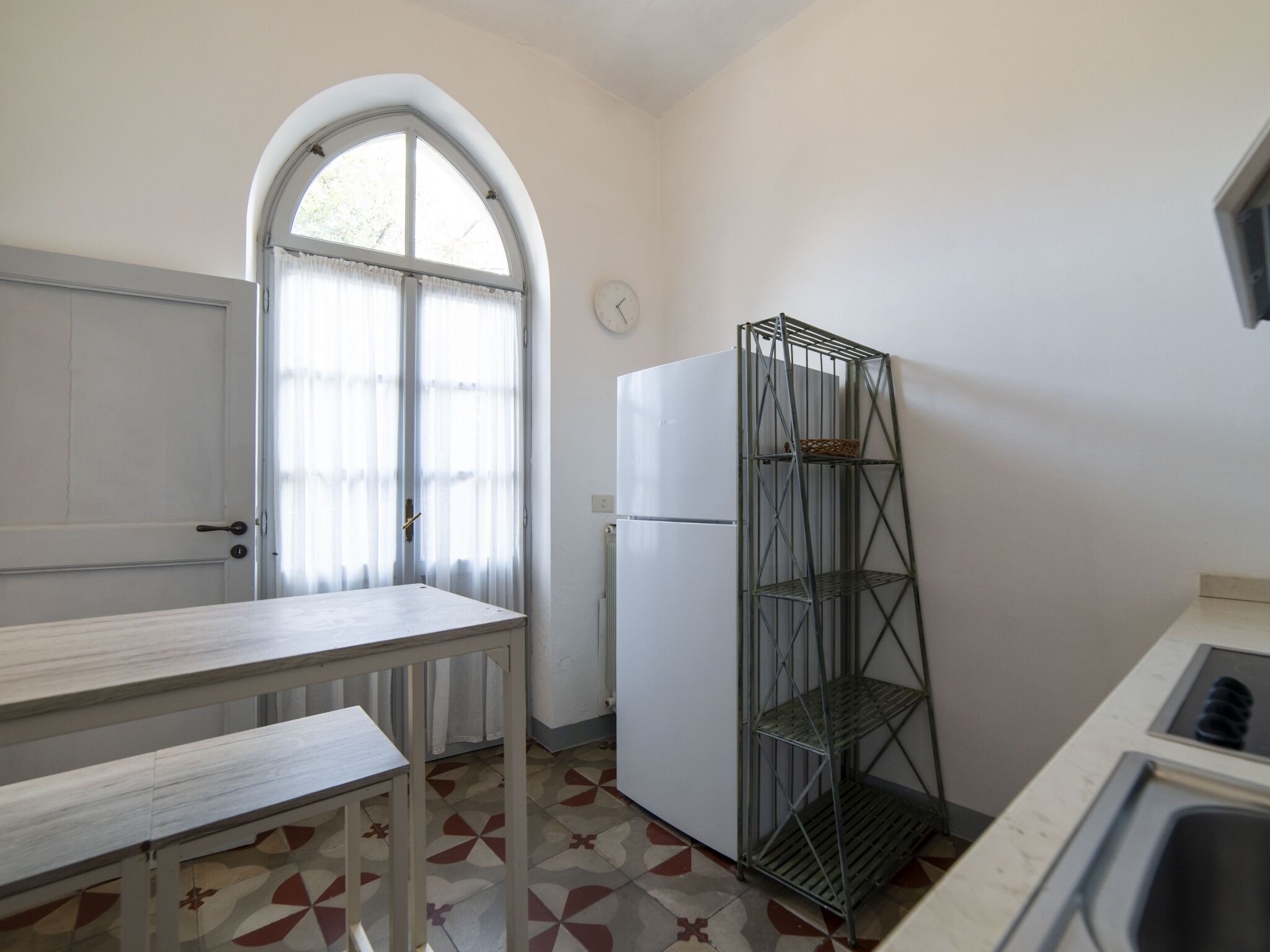 Una cuina petita amb un frigorífic blanc, una estanteria de metall, una finestra amb cortines i un paviment de rajoles.