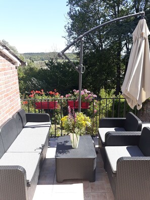 A patio with wicker furniture, a table with a vase of flowers, and a view of a garden with red flowers and trees. - L'escale Arquaise, La maison au jardin fleuri 5 km from dieppe 7 days minimum  (Arques-la-Bataille)